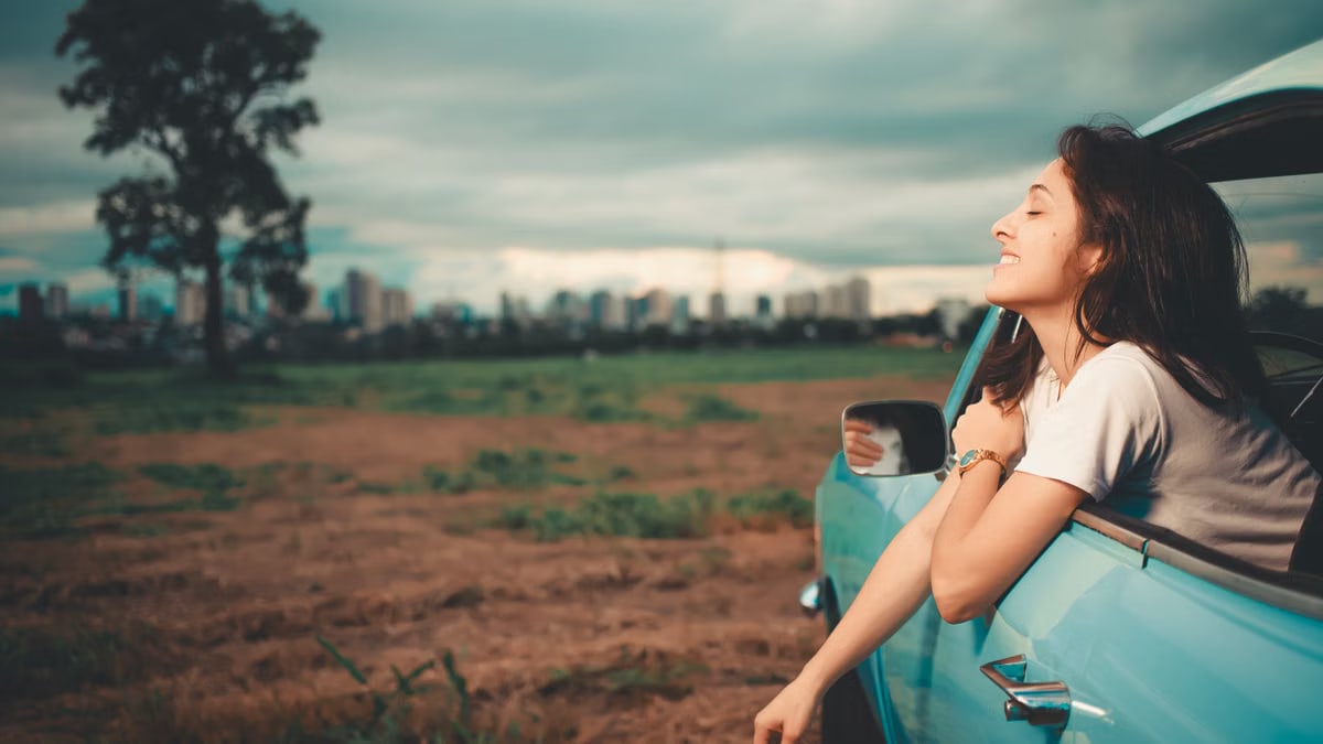 A young woman leans out of a blue car window, enjoying the scenic view during a summer road trip.